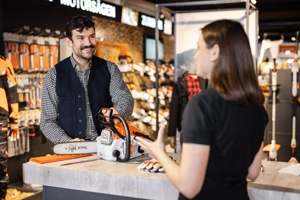 Un rivenditore STIHL consiglia una giovane donna sull'acquisto di un tagliasiepi STIHL davanti a uno scaffale di vendita