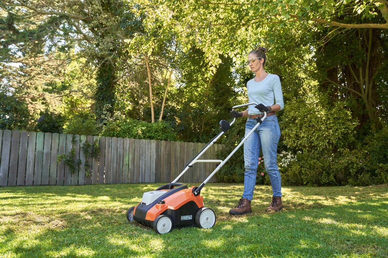 Donna che arieggia il prato in giardino con l’arieggiatore a batteria STIHL RLA 240, dotata di occhiali protettivi e guanti da lavoro.