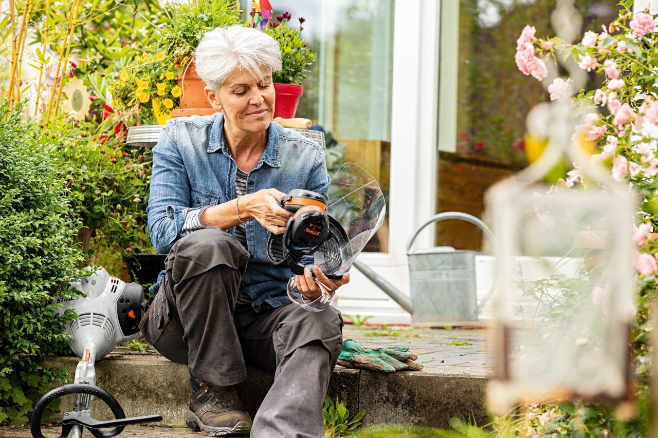 Donna seduta tra le piante in fiore accanto a un decespugliatore STIHL FS 38 che osserva i dispositivi di protezione individuale