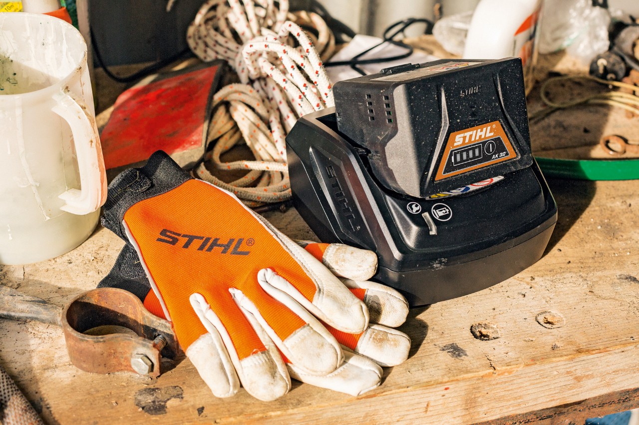 Batterie agli ioni di litio della serie AK in ricarica accanto a un paio di guanti e agli attrezzi da lavoro STIHL