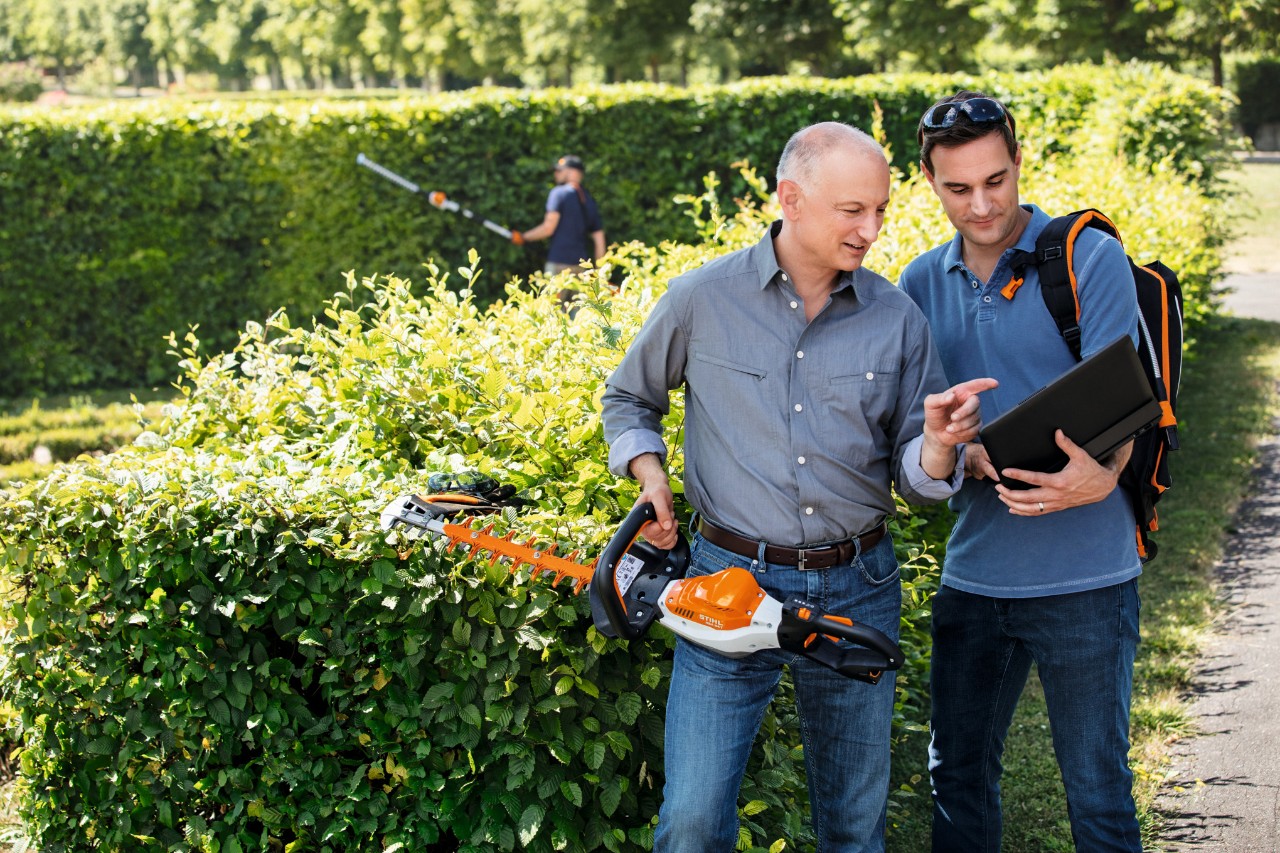 Il Dr. Nikolas Stihl tiene in mano un tagliasiepi STIHL e parla con una persona in piedi accanto a lui con un computer portatile.